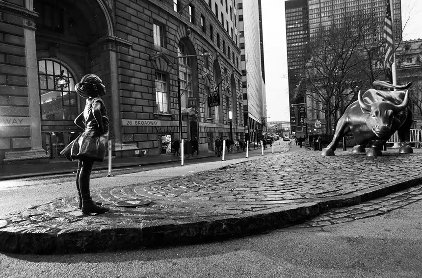 Fearless girl. Criança observando a escultura do touro da bolsa de valores em Wall Street, Nova York, na rua com prédios ao fundo, em uma foto em preto e branco.