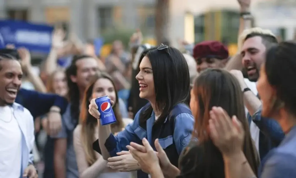 Kendall Jenner sorridente em uma celebração com um refrigerante Pepsi, cercada por um grupo de amigos alegres em ambiente de festa ao ar livre.