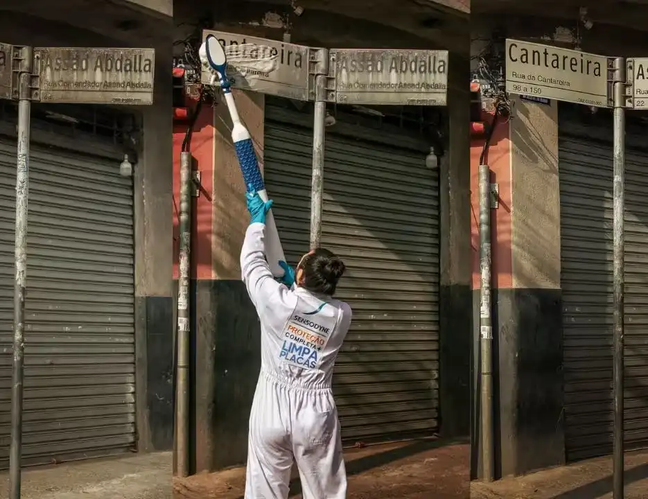 Campanha Sensodyne  "limpa placas. Funcionária de limpeza utilizando uma vassoura para limpar uma placa de sinalização em uma rua fechada, com prédios de fundo, durante a noite.