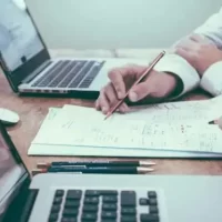 Duas pessoas trabalhando em uma mesa com laptops e anotações, discutindo um projeto juntos