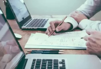 Duas pessoas trabalhando em uma mesa com laptops e anota&ccedil;&otilde;es, discutindo um projeto juntos