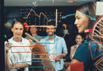 Equipe de profissionais discutindo estrat&eacute;gias de marketing digital, com gr&aacute;ficos e anota&ccedil;&otilde;es em uma tela de vidro, demonstrando planejamento e inova&ccedil;&atilde;o.