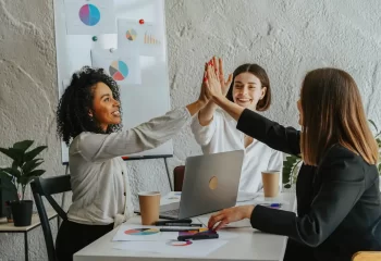 Tr&ecirc;s mulheres de escrit&oacute;rio comemorando uma conquista, s&iacute;mbolo de sucesso em estrat&eacute;gias de go-to-market, em ambiente de trabalho colaborativo com gr&aacute;ficos.