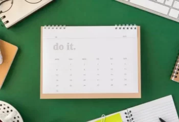 Flat lay de mesa com fundo verde, agenda, calend&aacute;rio com "do it", &oacute;culos, teclado, canetas, ch&aacute;, despertador e blocos de notas