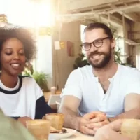 Grupo de amigos sorrindo e conversando em um café, rodeados por plantas. A imagem retrata um ambiente descontraído e social.