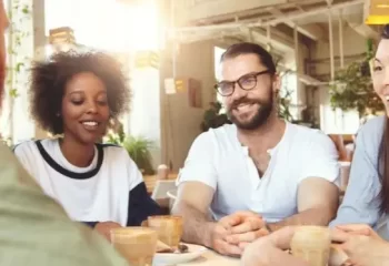 Grupo de amigos sorrindo e conversando em um caf&eacute;, rodeados por plantas. A imagem retrata um ambiente descontra&iacute;do e social.