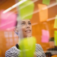 Mulher pensando e planejando usando post-its coloridos em um quadro de planejamento ou brainstorm, simbolizando organização e criatividade.