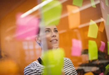 Mulher pensando e planejando usando post-its coloridos em um quadro de planejamento ou brainstorm, simbolizando organização e criatividade.