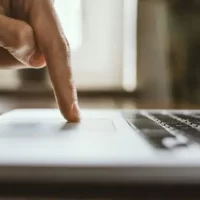 Imagem de uma mão pressionando o touchpad de um laptop, simbolizando o uso de tecnologia e interatividade digital em ambientes de trabalho e estudo.