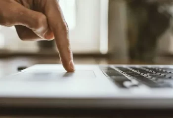 Imagem de uma m&atilde;o pressionando o touchpad de um laptop, simbolizando o uso de tecnologia e interatividade digital em ambientes de trabalho e estudo.
