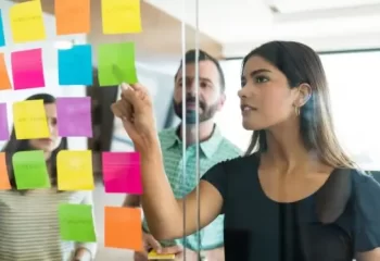 Mulher discutindo post-it com colegas de trabalho em reuni&atilde;o no escrit&oacute;rio