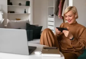 Mulher sorridente jogando no smartphone enquanto est&aacute; sentada em um ambiente confort&aacute;vel com um laptop e itens de trabalho ao redor.
