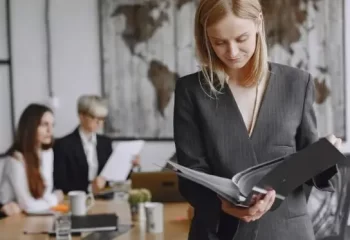 Uma mulher em trajes formais revisando documentos em uma sala de reuni&otilde;es, com mapas ao fundo e outras pessoas trabalhando.