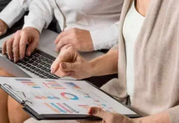 Mulher sentada apontando para gr&aacute;ficos em prancheta nas m&atilde;os, junto &agrave; colega de trabalho com notebook no colo