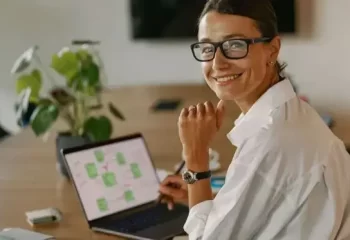 Mulher sorridente usando &oacute;culos em ambiente de trabalho moderno, olhando para a c&acirc;mera enquanto trabalha em laptop sobre mesa de madeira.