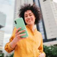 Mulher sorrindo usando camisa amarela segurando smartphone com capa verde em área urbana com prédios ao fundo