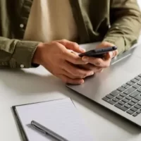 Pessoa usando smartphone, com notebook e bloco de anotações sobre a mesa