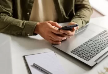 Pessoa usando smartphone, com notebook e bloco de anota&ccedil;&otilde;es sobre a mesa